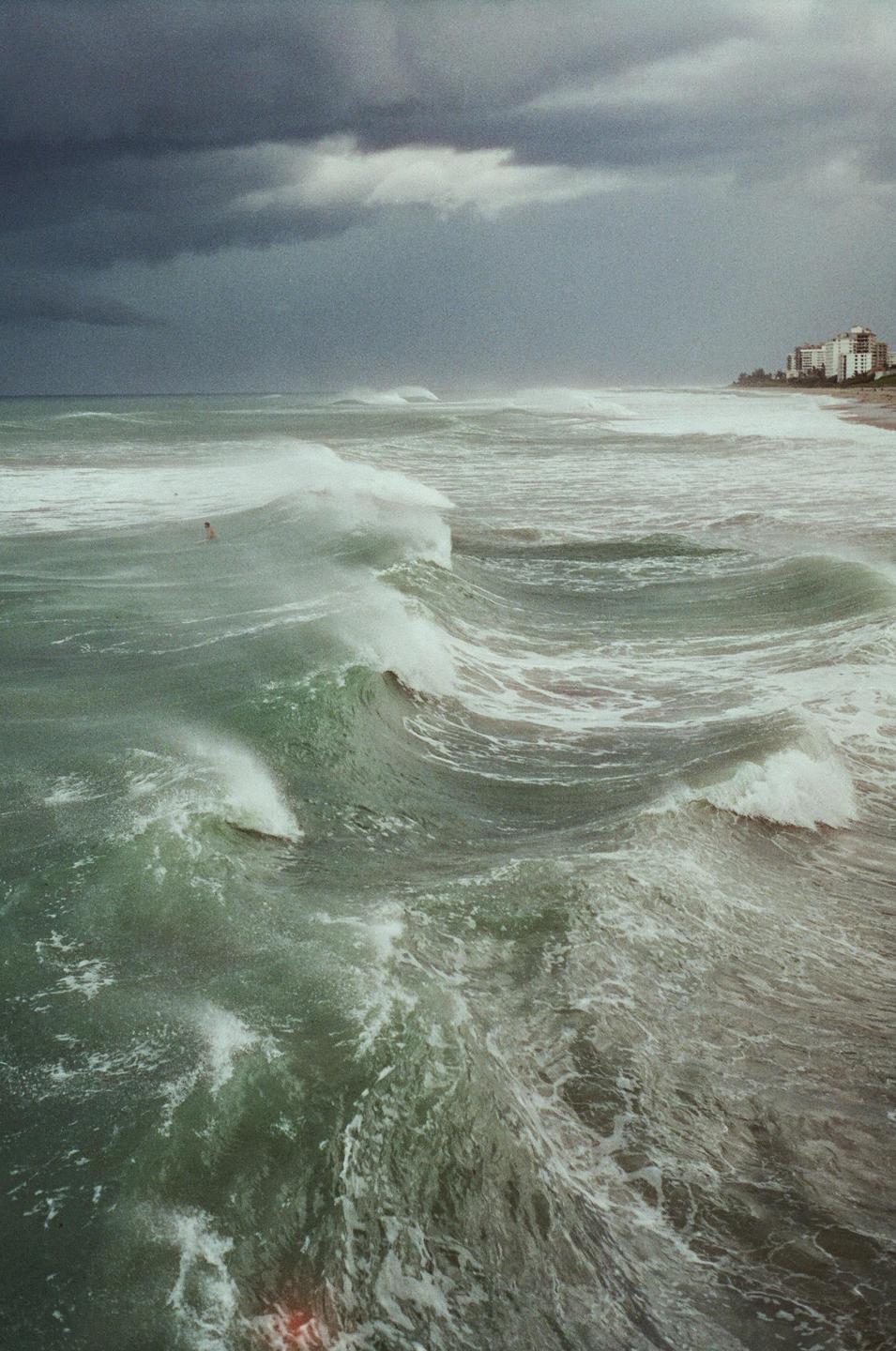 Large waves crash on a stormy ocean with buildings visible on the distant shore.