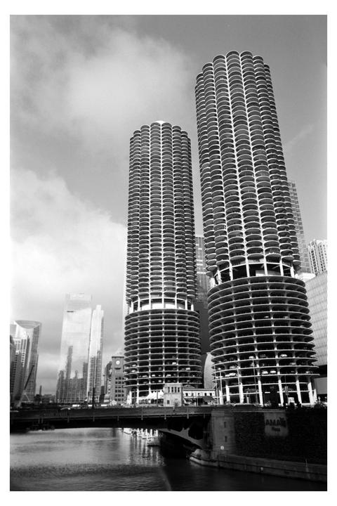 Two cylindrical high-rise buildings with many balconies stand next to a river with a bridge over it.