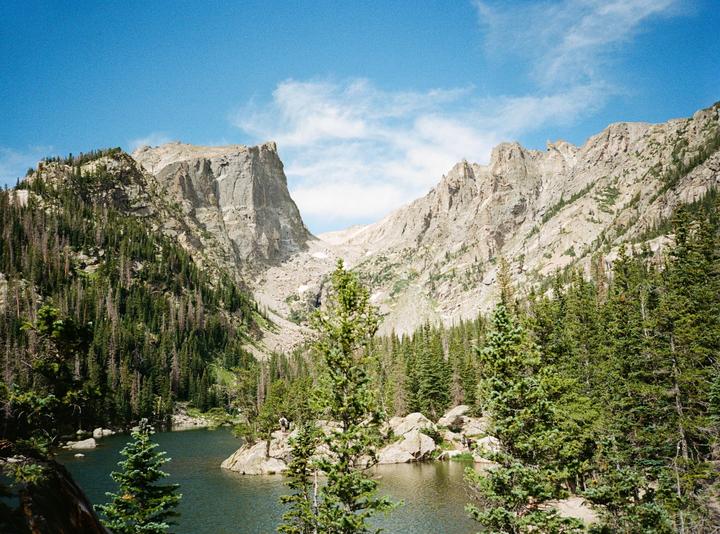 A lake is surrounded by a dense forest and rocky mountains under a blue sky with scattered clouds.