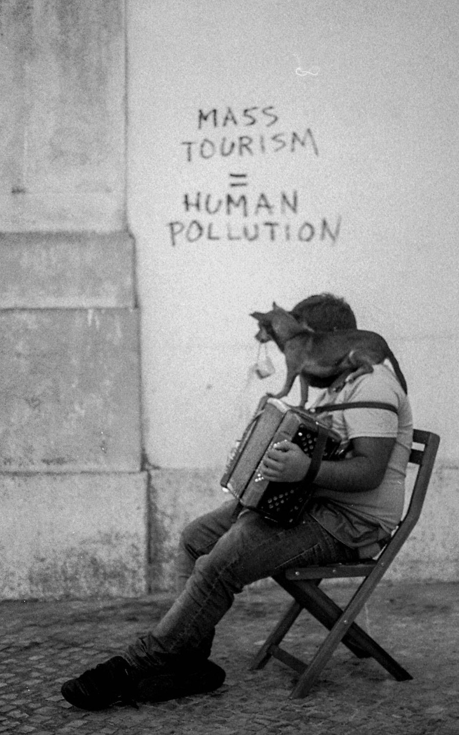 A man sits on a folding chair playing an accordion with a small dog on his shoulder, next to a wall with graffiti that reads "MASS TOURISM = HUMAN POLLUTION."