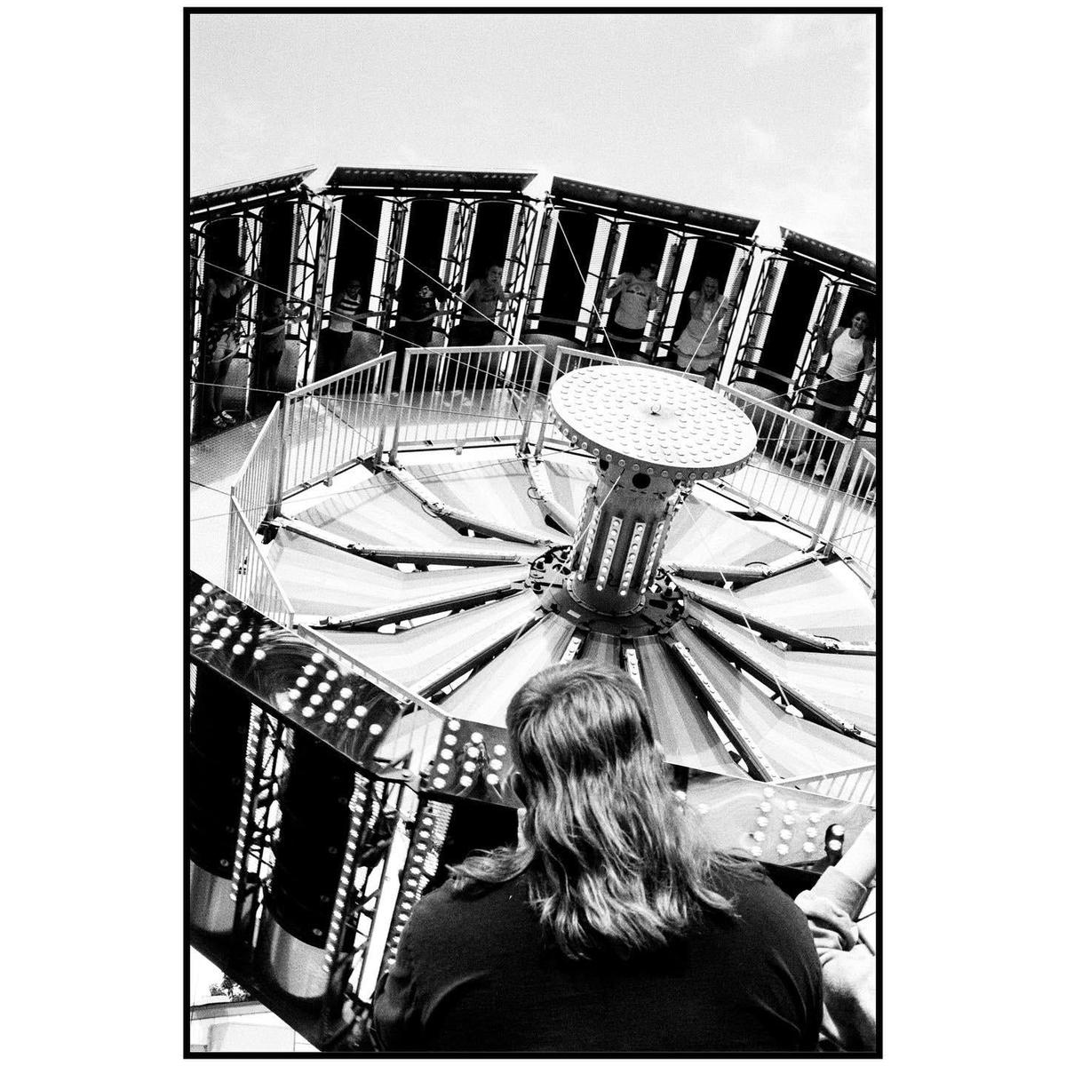 A person with long hair sits in the foreground of a spinning carnival ride with other people visible in the background.