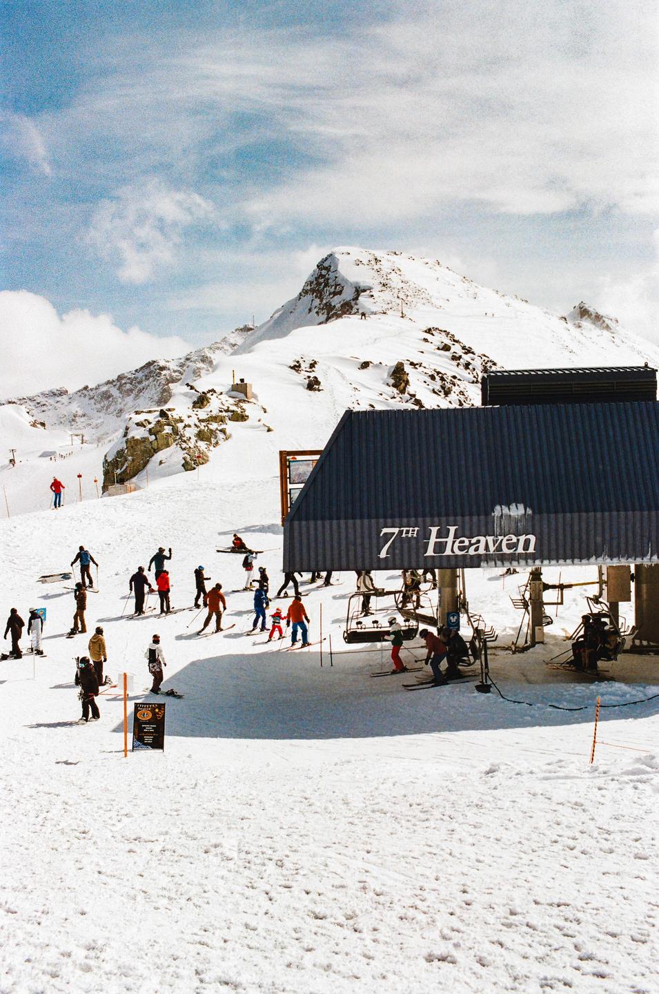 A group of skiers and snowboarders gather at the base of a ski lift named "7th Heaven" on a snowy mountain.