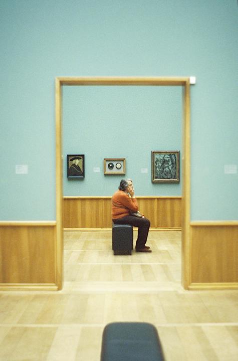 A man sits on a stool in a museum gallery with paintings on the wall.