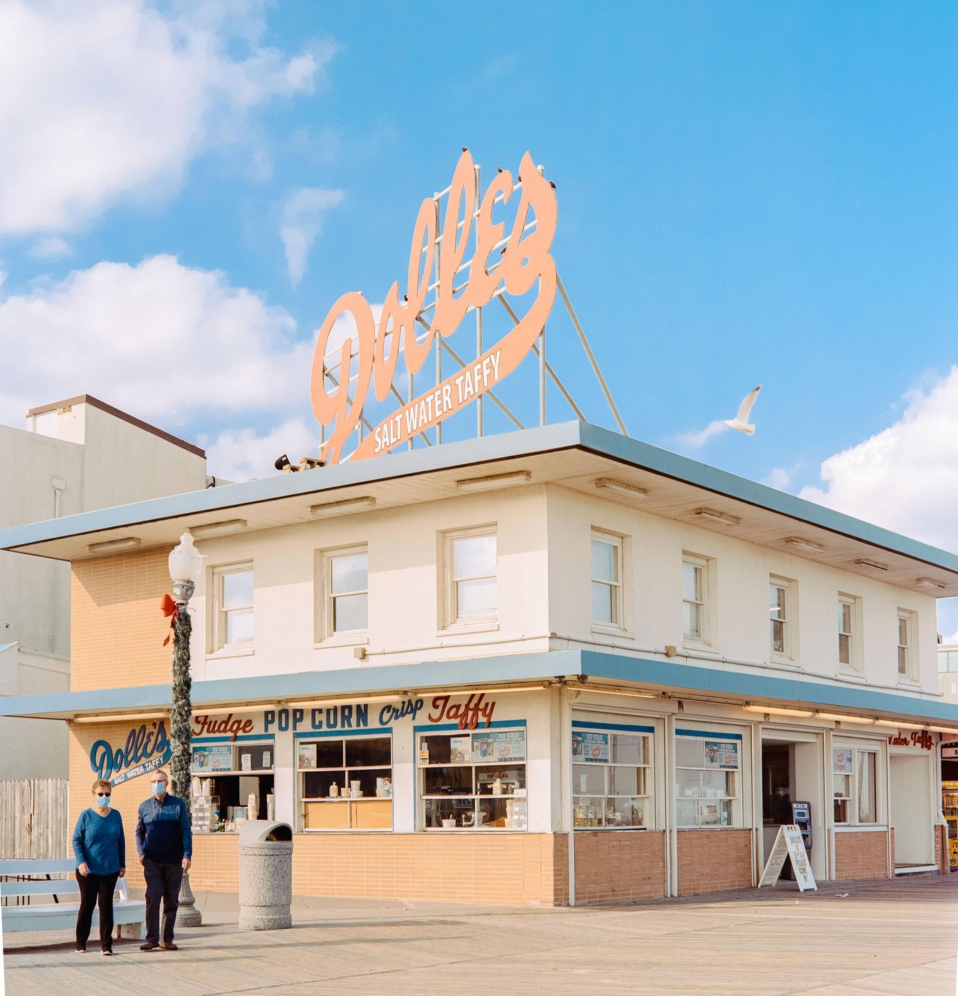 Two people stand on a boardwalk in front of a building with a large sign for Dollie's Salt Water Taffy.