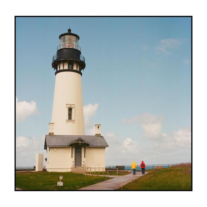 Two people walk away from a white lighthouse with a black top under a blue sky with scattered clouds.