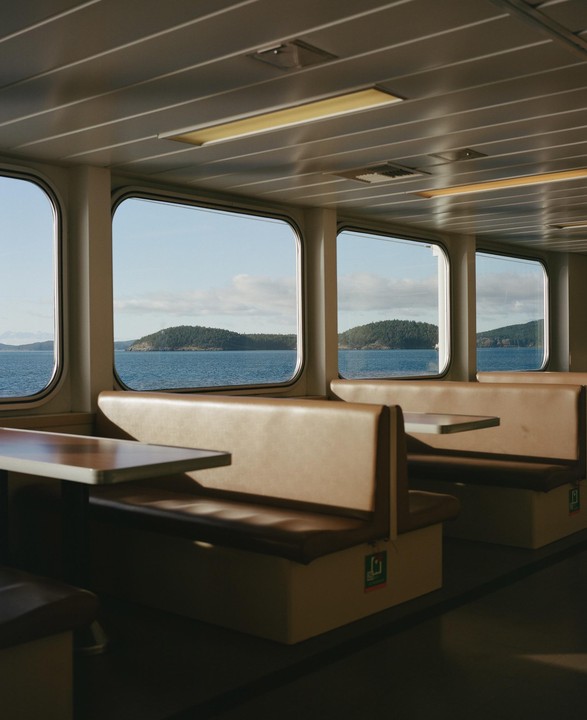 The interior of a ferry shows rows of empty booths with windows looking out onto a body of water with tree-covered islands.