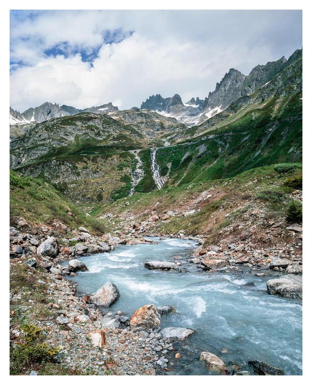 A river flows through a rocky valley with mountains and a waterfall in the background.