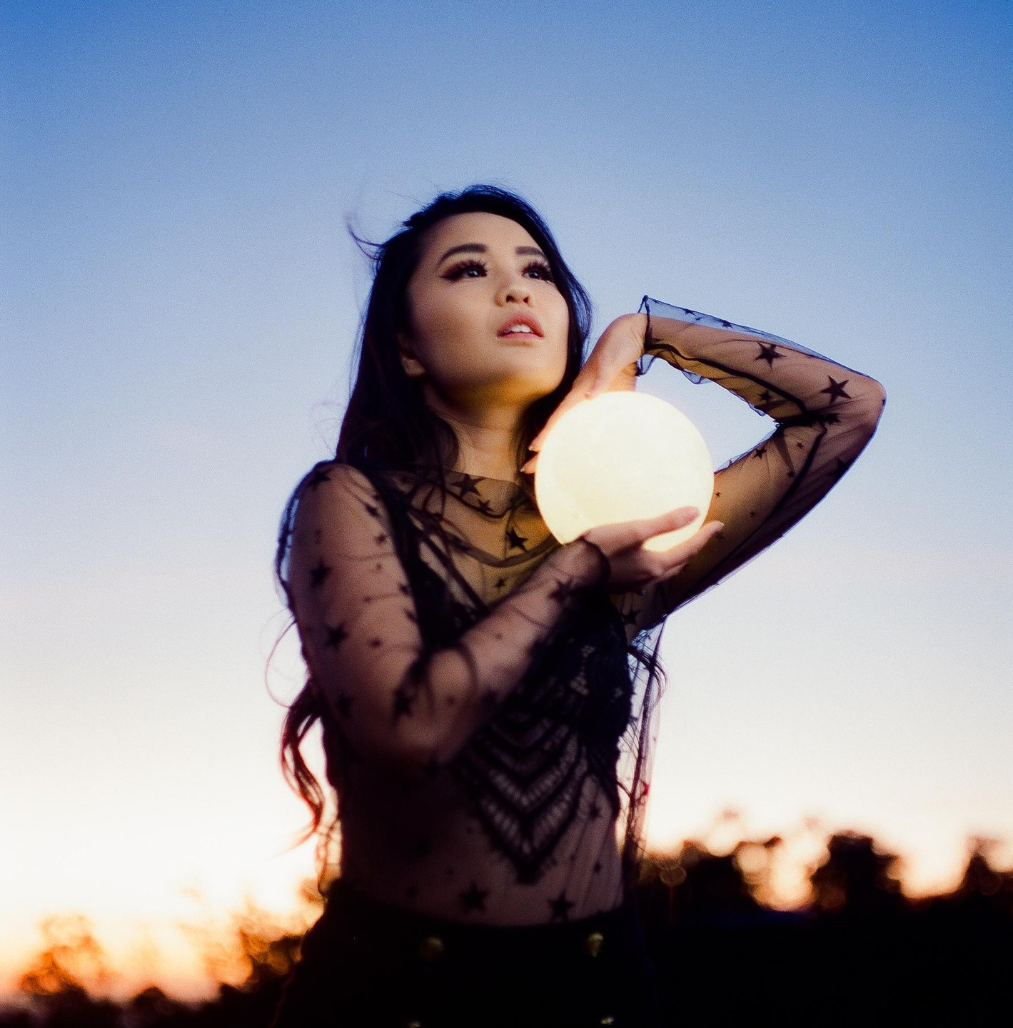 A woman in a sheer top with stars holds a glowing orb against a twilight sky.