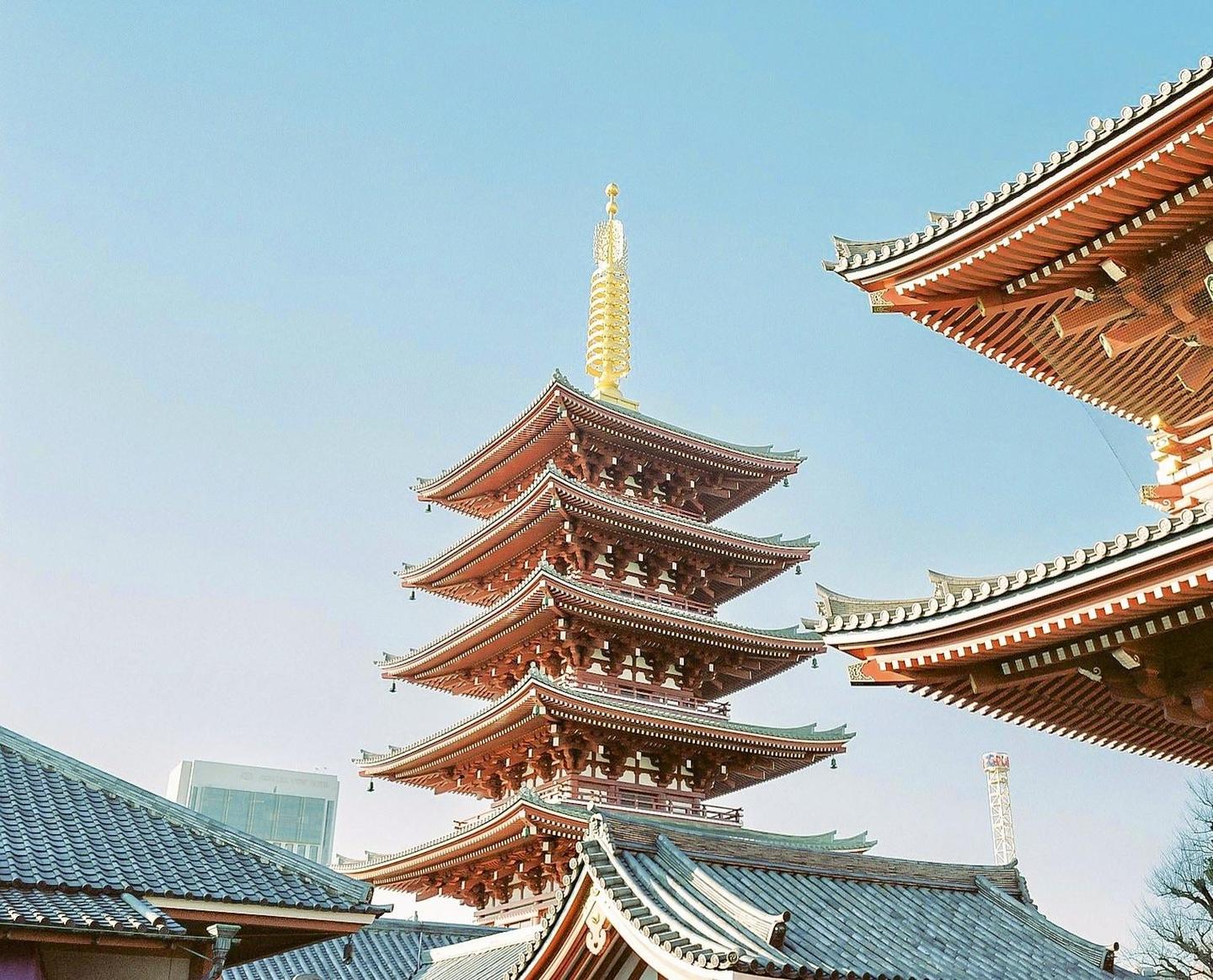 A red pagoda with a golden spire stands tall against a clear blue sky, surrounded by other traditional Japanese rooftops.