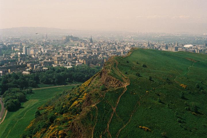 A city with a castle on a hill is visible behind a large green hill with walking paths.
