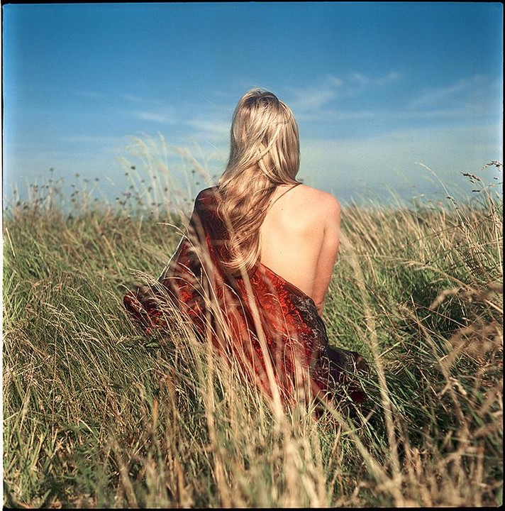 A woman with long blonde hair sits in a field of tall grass, her back to the viewer, wearing a red garment.
