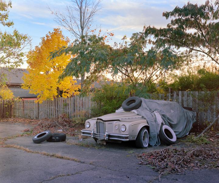 An abandoned car covered with a tarp and tires on top is parked next to a wooden fence with autumn trees in the background.