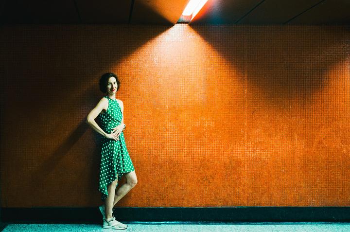 A woman in a green polka dot dress stands against an orange tiled wall with a bright light above her.