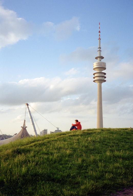 A man sits on a grassy hill with a tall telecommunications tower and a large tent-like structure in the background.