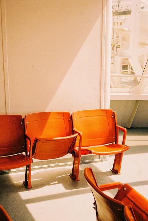 Three orange seats are arranged in a row on a ferry deck, with sunlight casting shadows on the floor and wall.