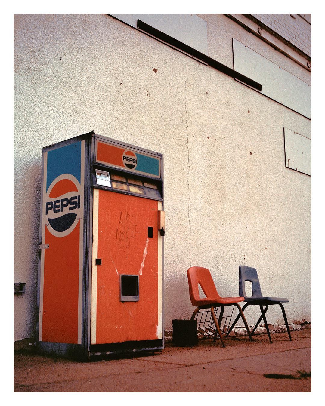 An old Pepsi vending machine stands next to two chairs against a white wall.