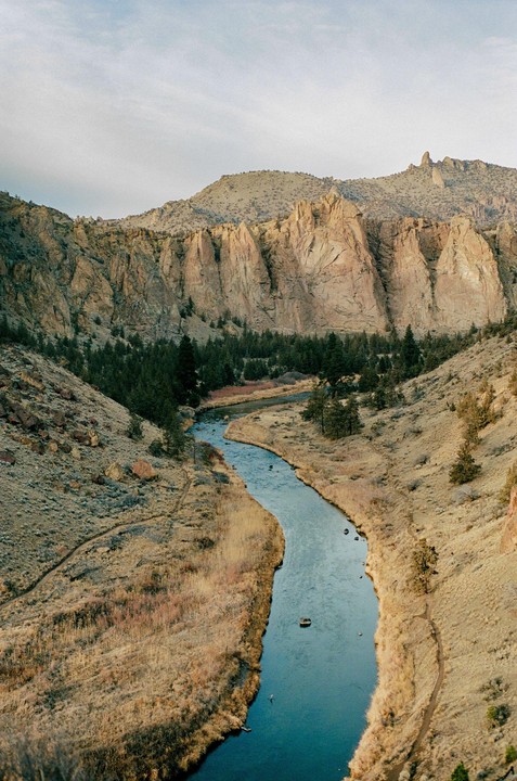 A river winds through a canyon with steep, rocky cliffs and scattered trees under a pale sky.