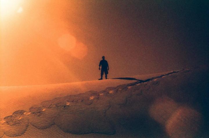 A man stands silhouetted on a sand dune under an orange sky.