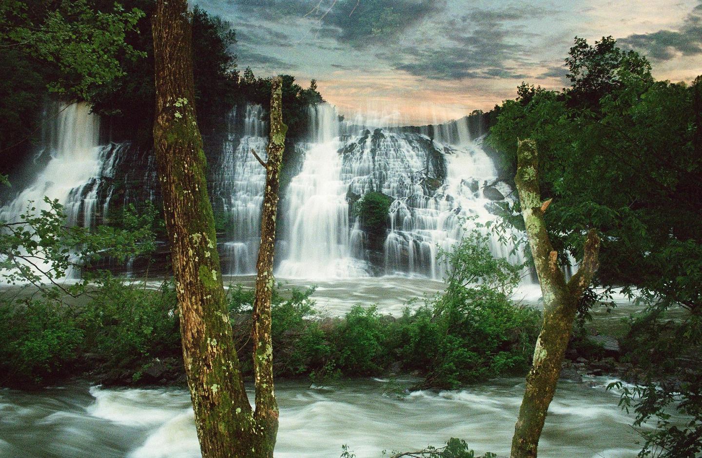 A large waterfall cascades into a river, framed by trees and lush foliage under a cloudy sky.