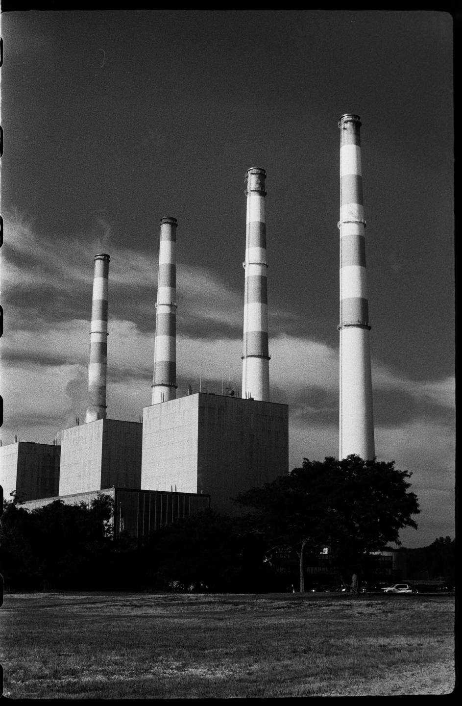 Four striped smokestacks rise above a power plant with a grassy field and trees in the foreground.