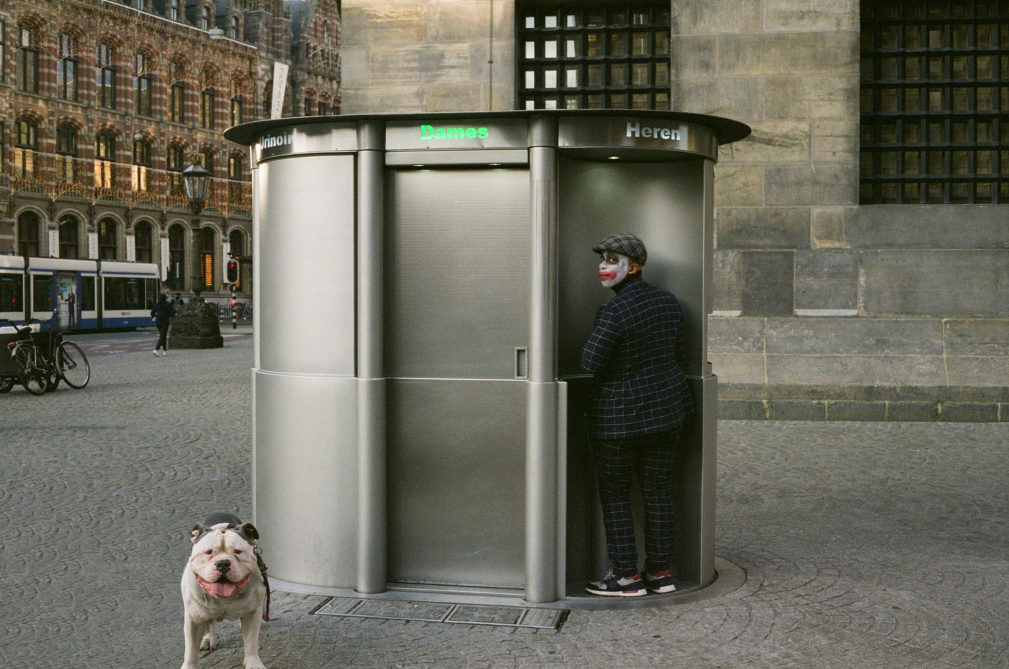 A man in clown makeup and a plaid suit stands at a public urinal while a bulldog looks at the camera on a cobblestone street in Amsterdam.