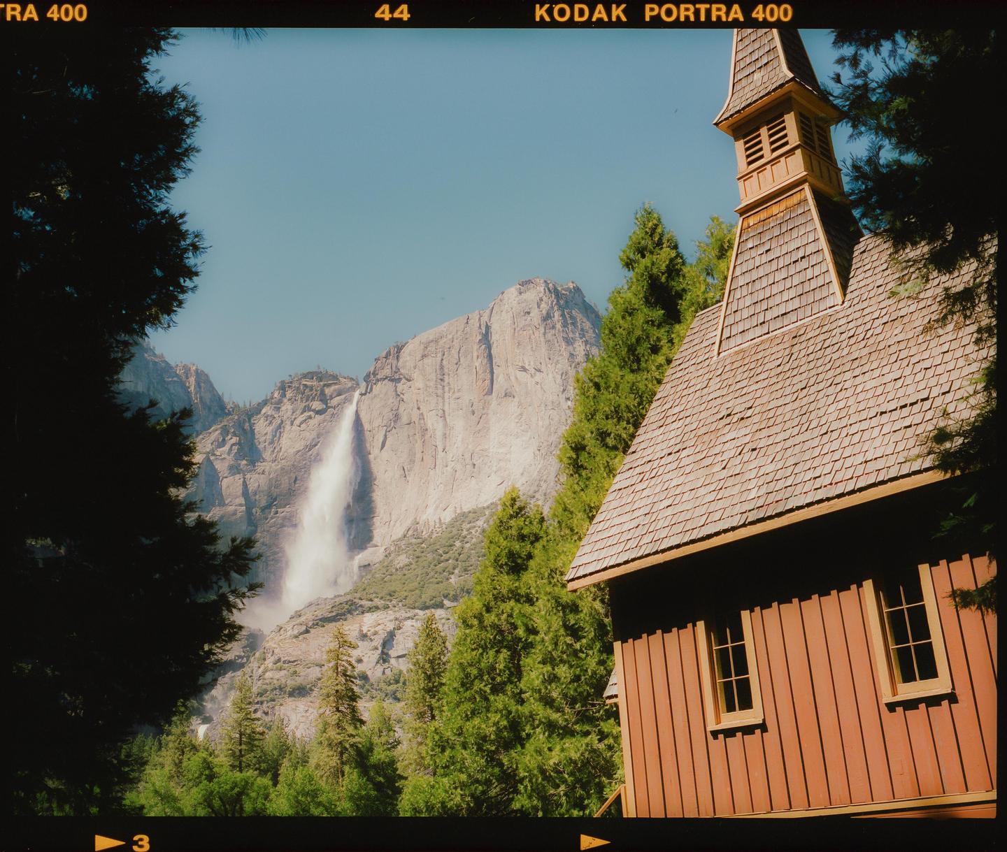 A wooden chapel with a tall steeple stands among trees, with a large waterfall and mountain in the background.