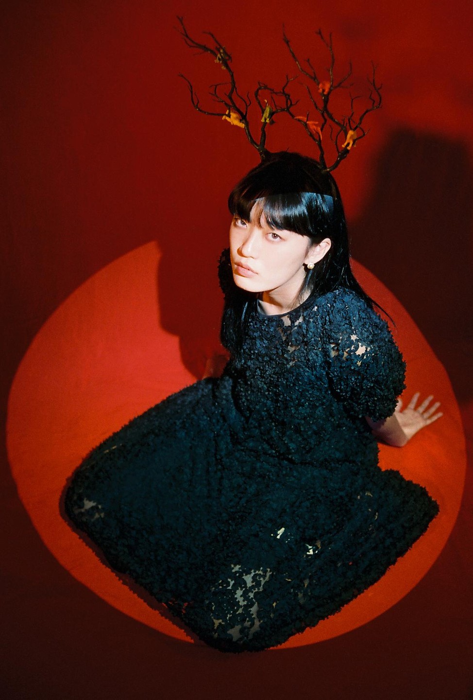 A woman wearing a black dress and a branch-like headpiece sits in a spotlight on a red background.