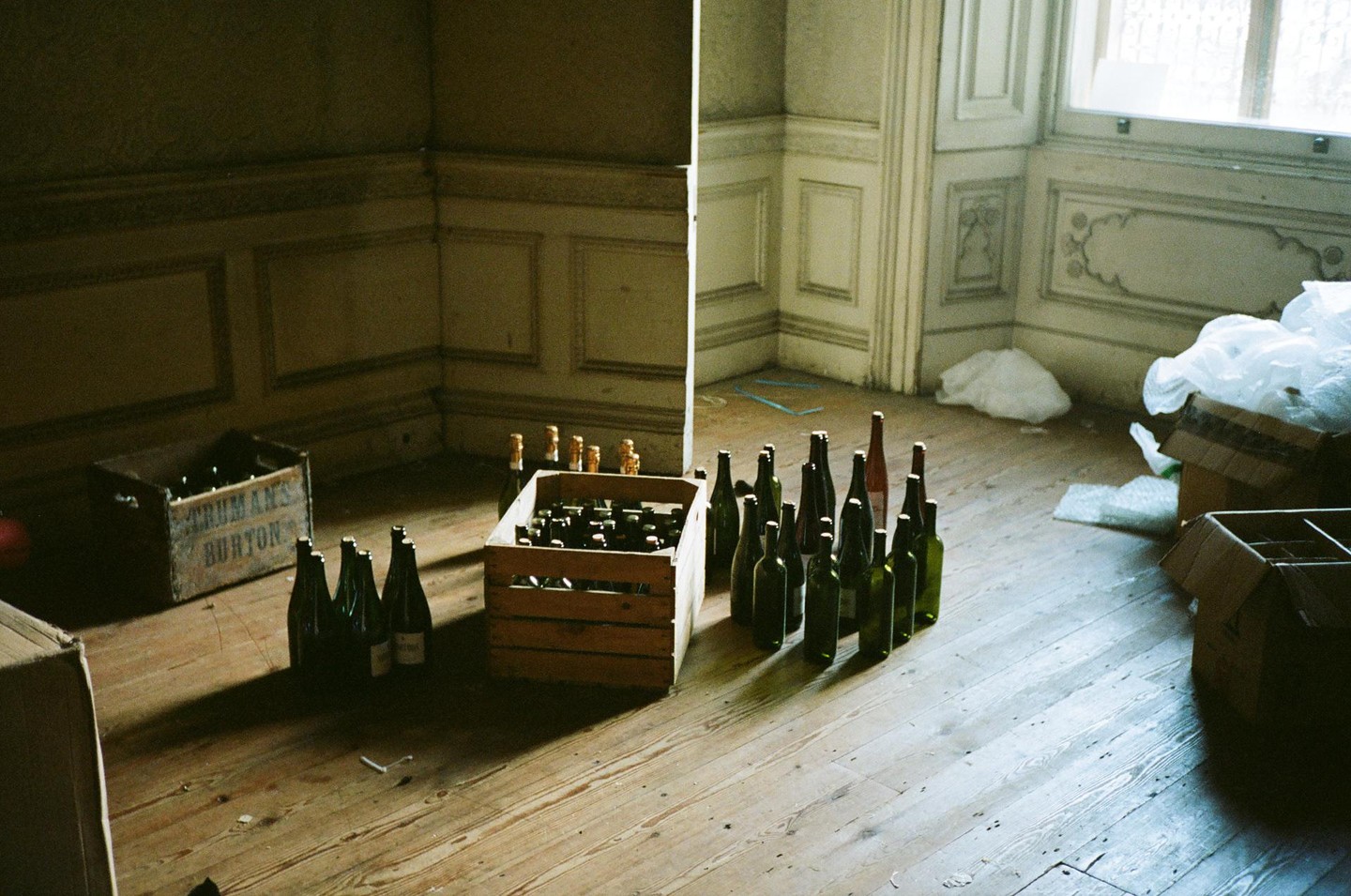 Empty bottles and wooden crates are scattered across a wooden floor in a room with ornate wall molding.