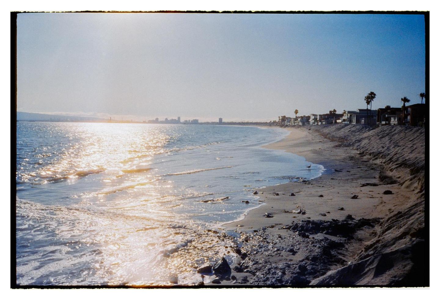The sun reflects brightly on the ocean waves along a sandy beach with houses and a city skyline in the distance.