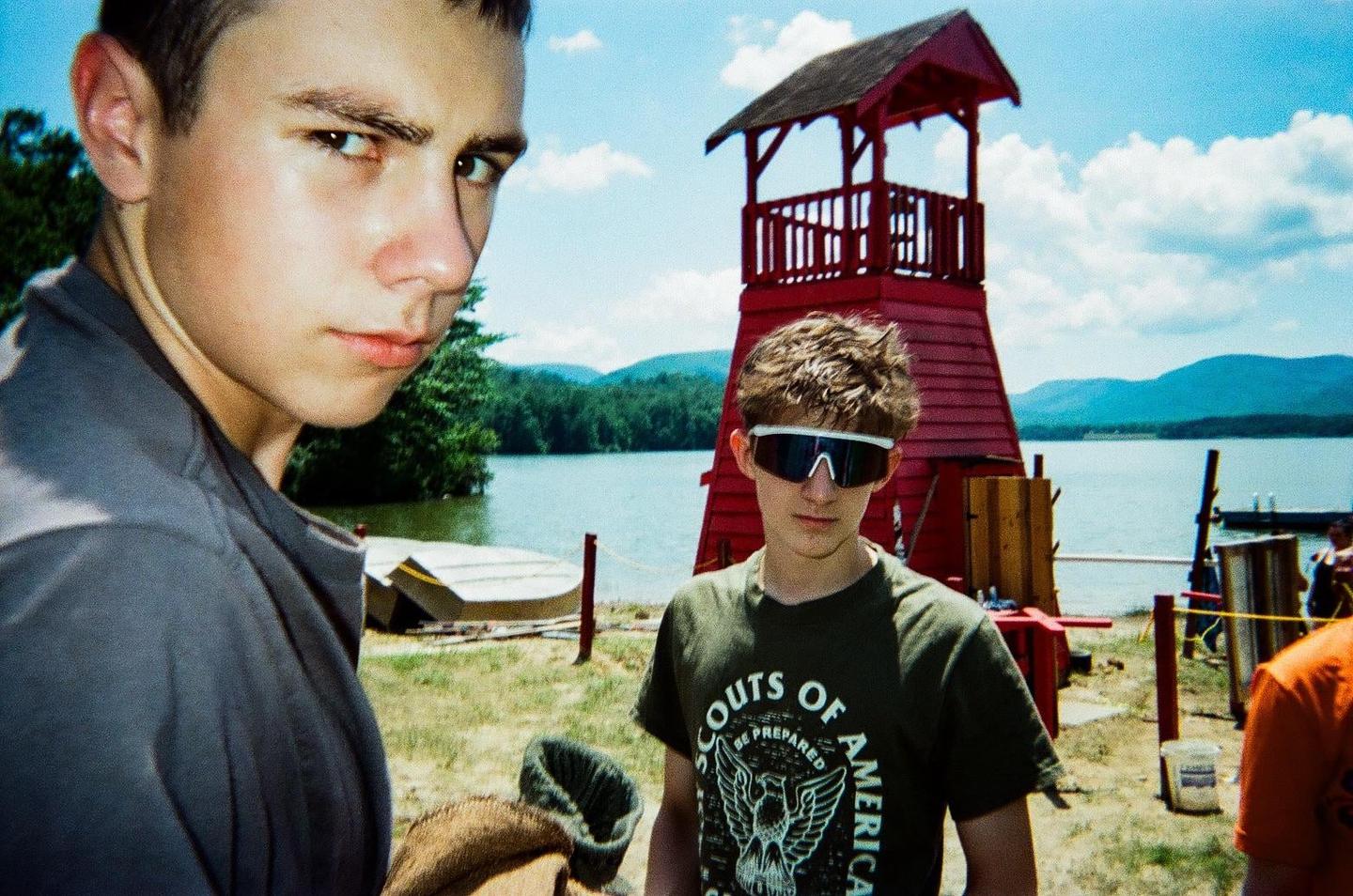Two boys stand near a lake with a red tower and mountains in the background.