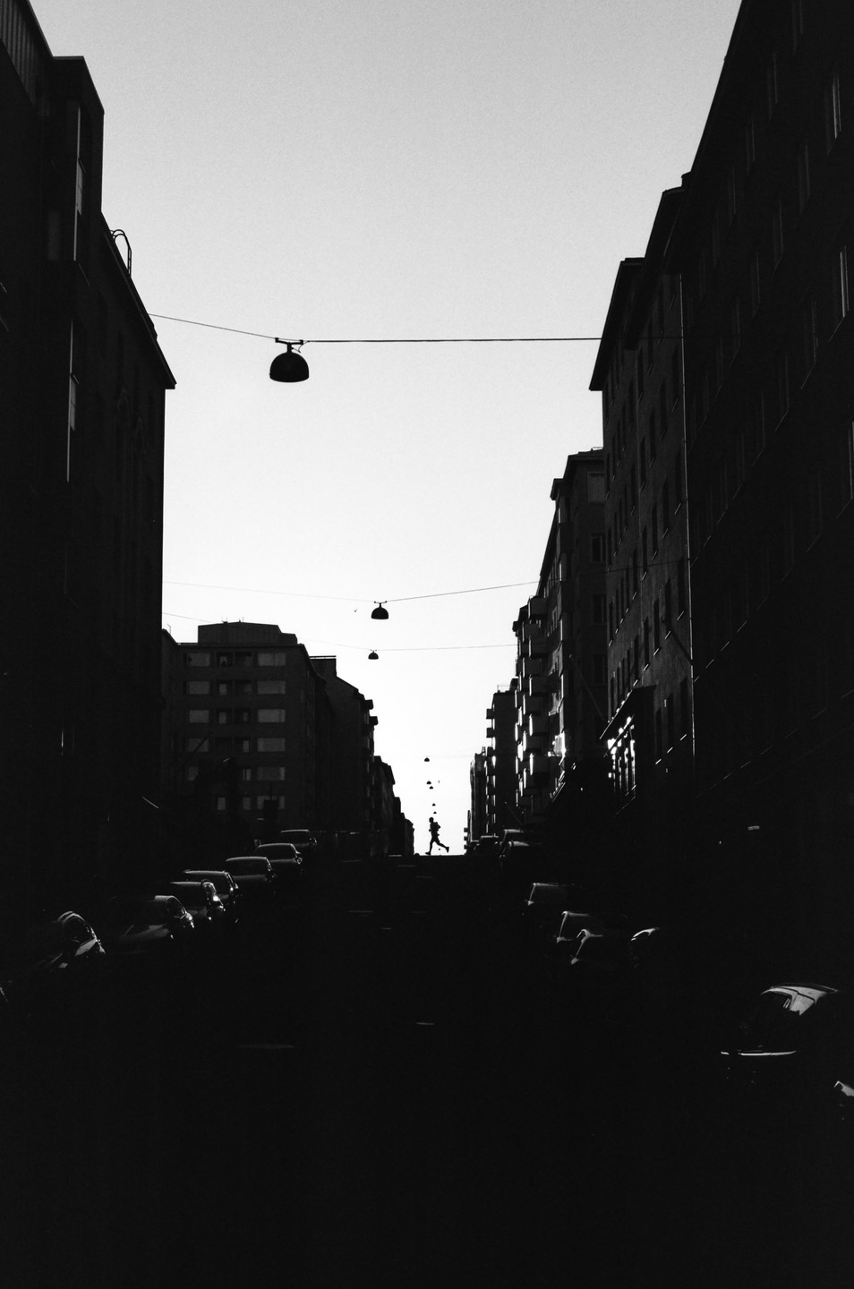 A person runs up a street lined with parked cars and tall buildings under a bright sky.