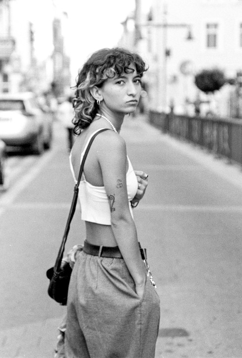 A woman with curly hair and a tattoo on her arm looks over her shoulder while standing on a street.