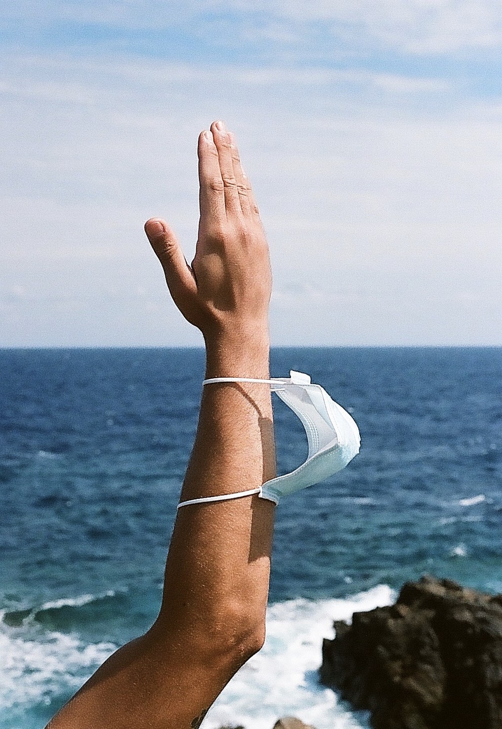 A person's arm is raised with a face mask hanging from the wrist against a backdrop of the ocean and sky.