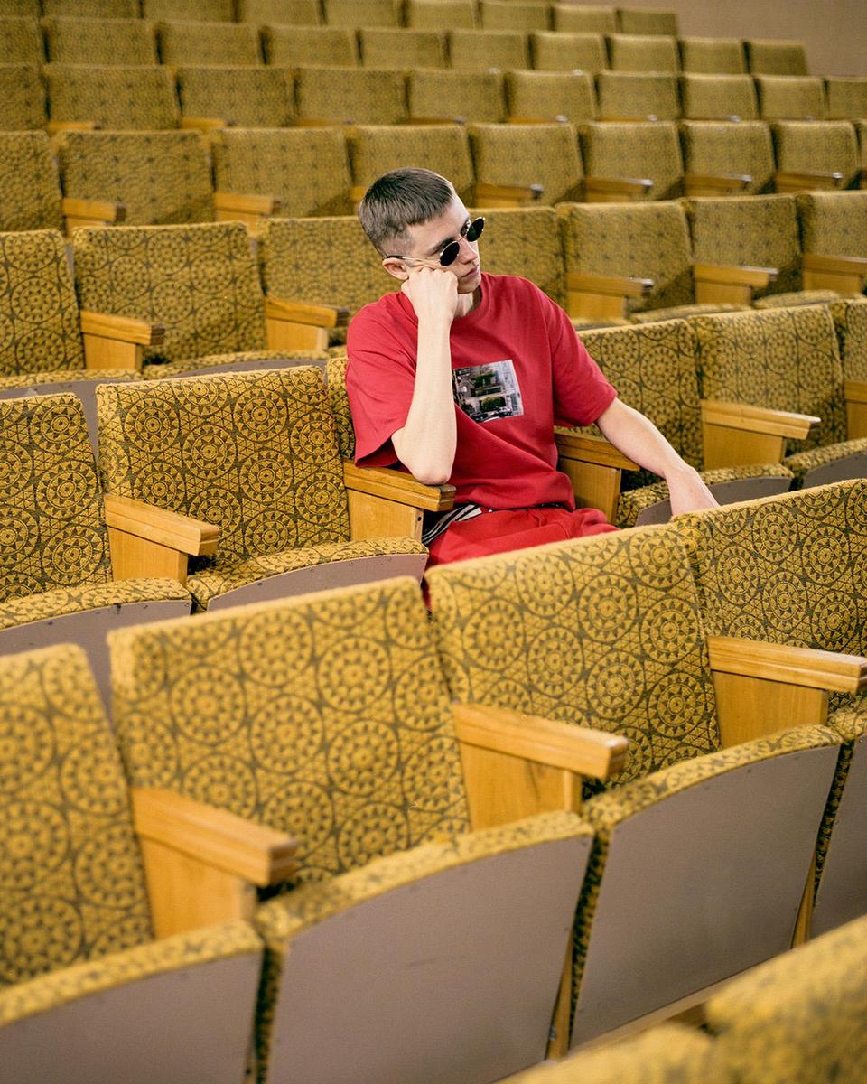 A man wearing sunglasses and a red shirt sits alone in an auditorium.