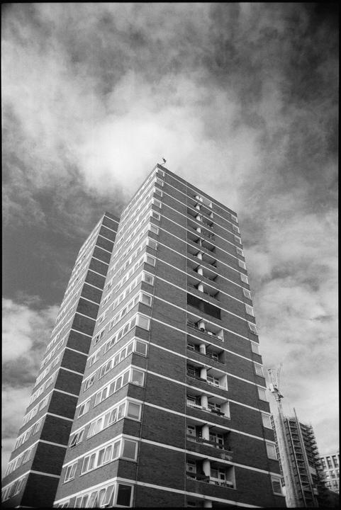 A low-angle black and white shot captures a tall brick building with many windows and balconies, with a cloudy sky above.