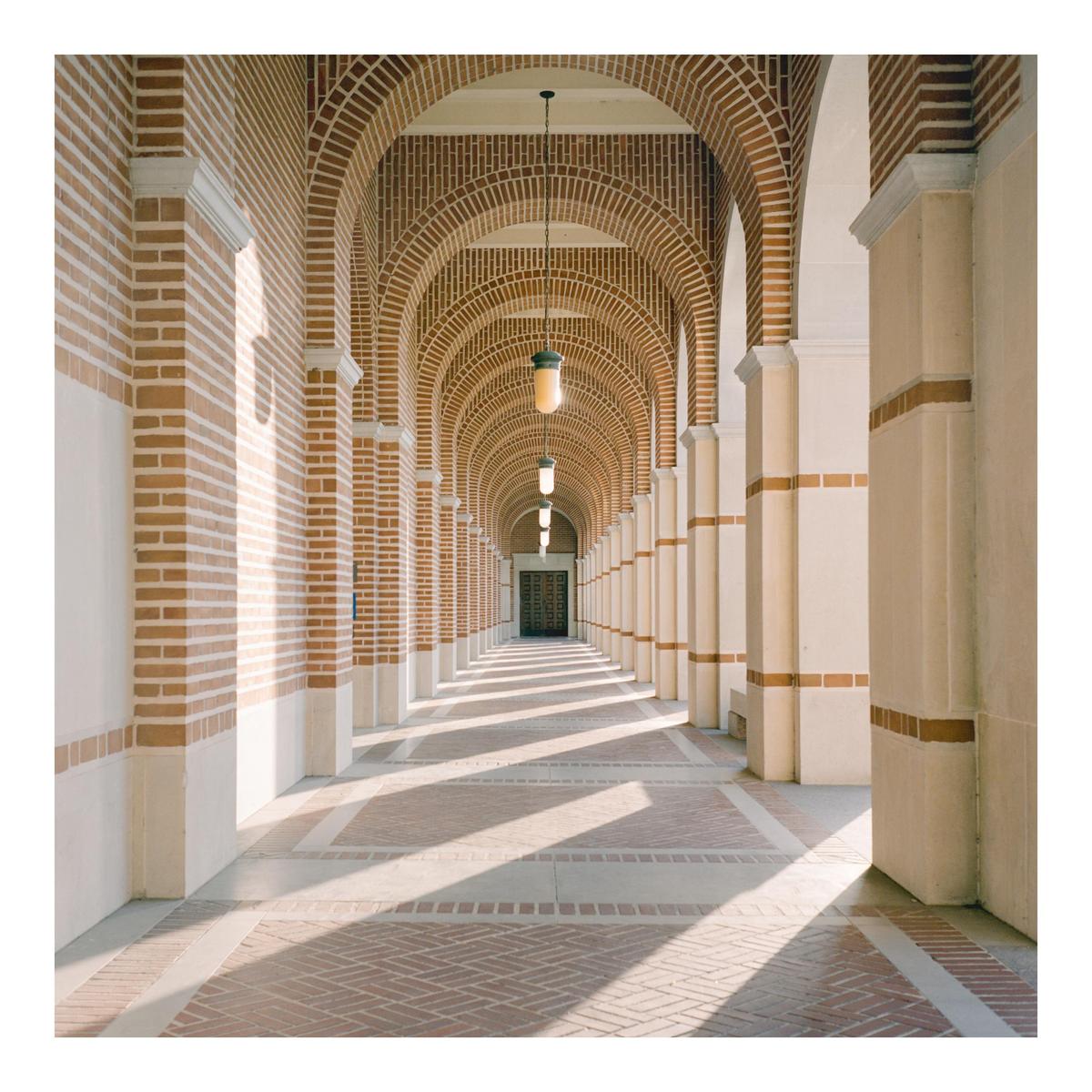 A long hallway with brick arches and columns is illuminated by sunlight creating shadows on the floor.