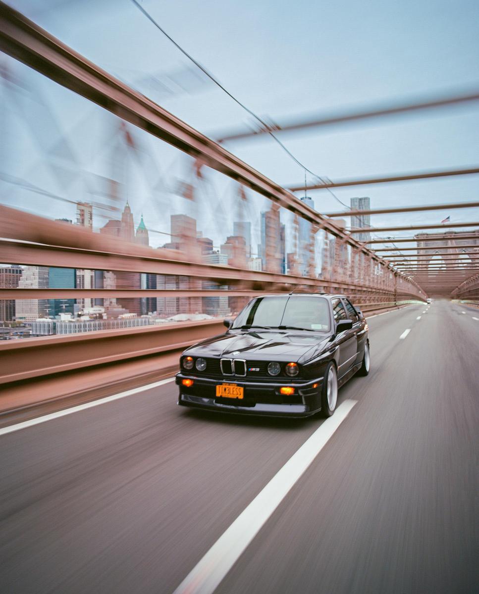 A black car drives on a bridge with a city skyline in the background.