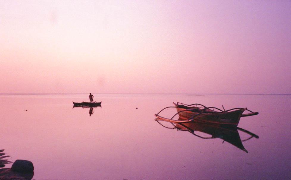 A man stands in a small boat on calm water with two other boats floating nearby at dusk.