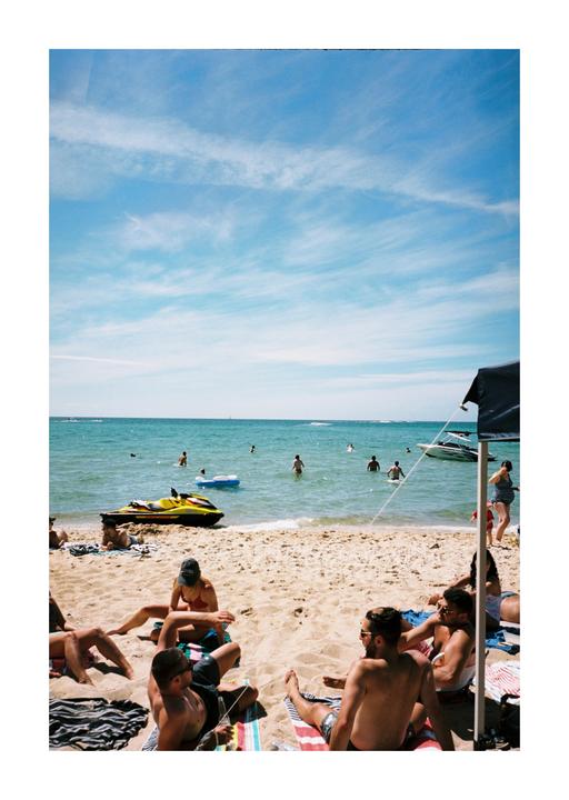 People are relaxing on a sandy beach with a jet ski and boats in the water under a blue sky.