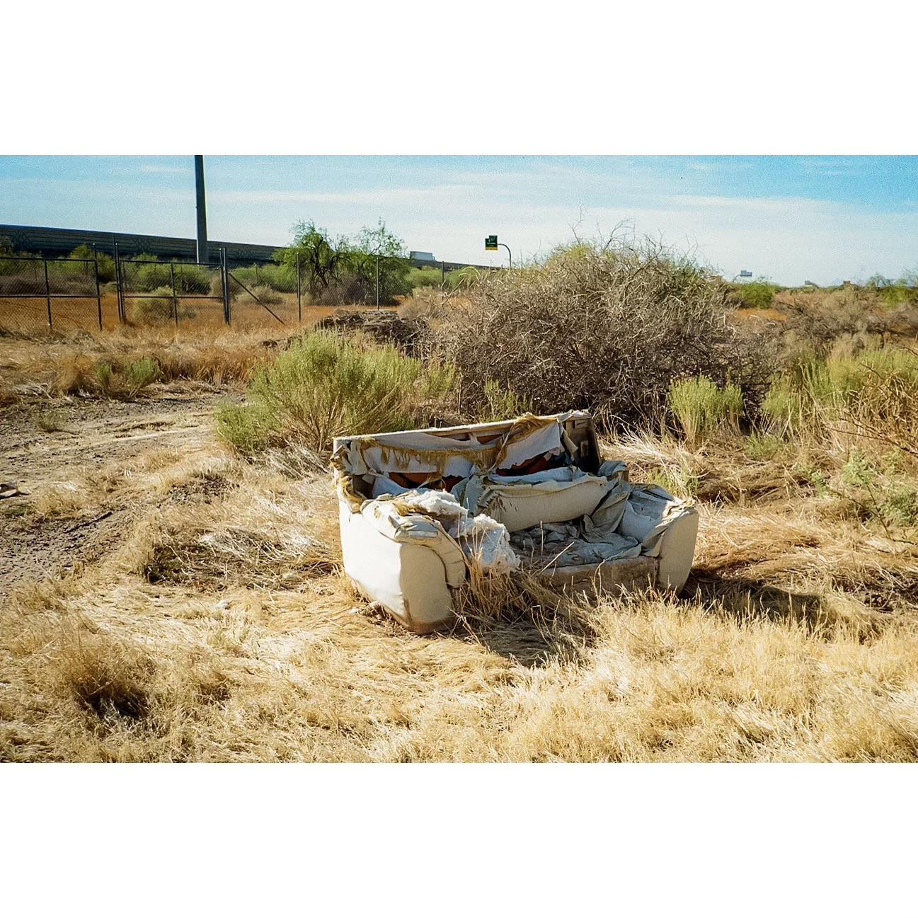 A tattered armchair sits in a field of dry grass and brush next to a highway.