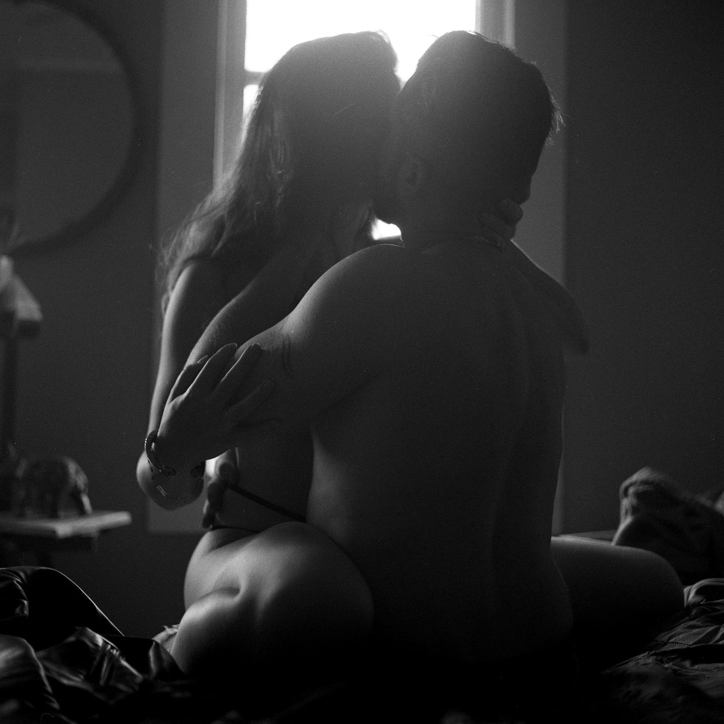 A couple kisses while sitting on a bed in a backlit room.