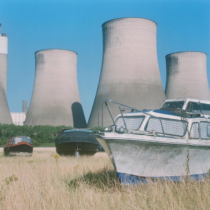 Three boats sit in a field of tall grass with several cooling towers in the background.