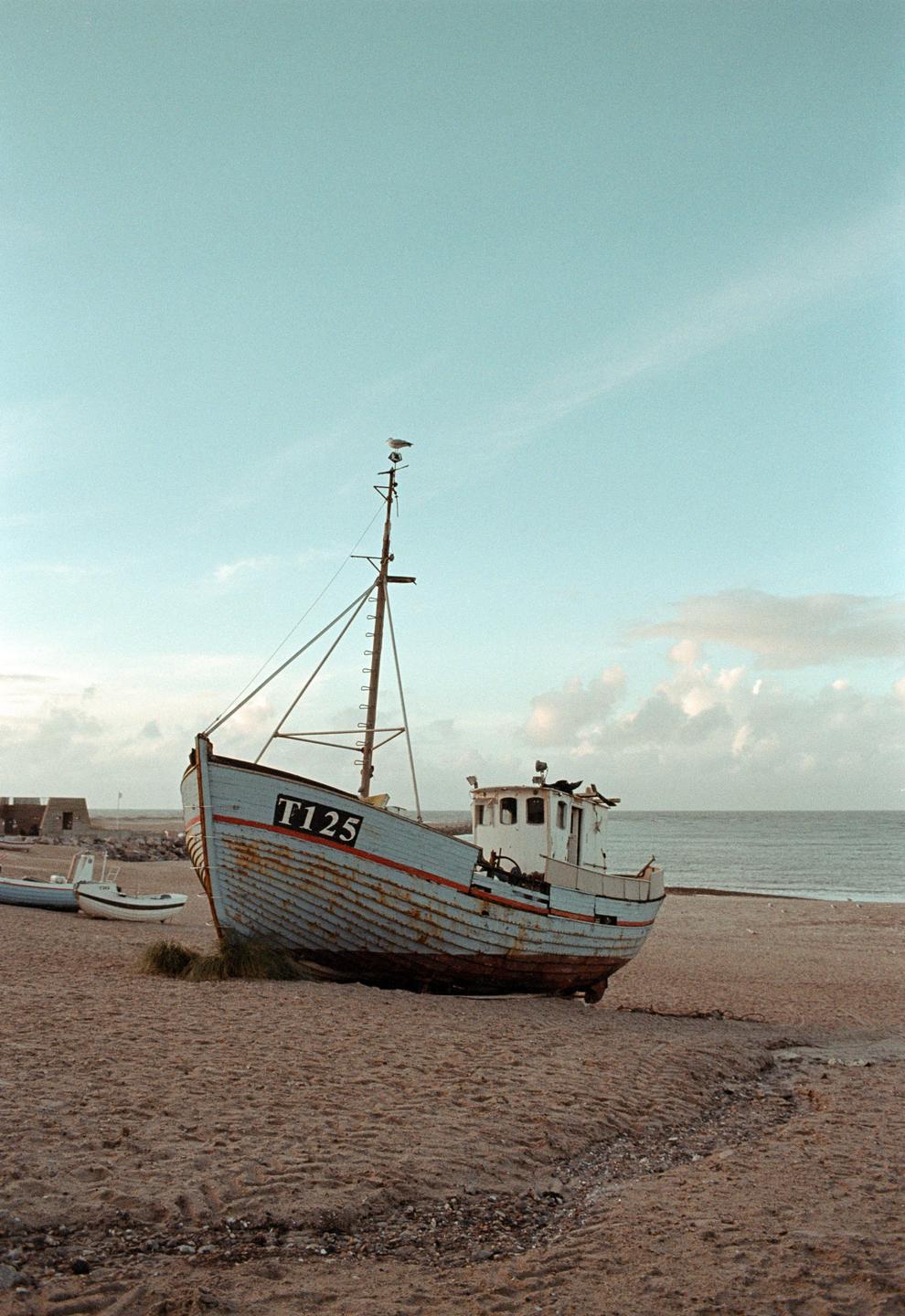 A fishing boat with the number T125 on its hull sits on a sandy beach next to the ocean under a cloudy sky.