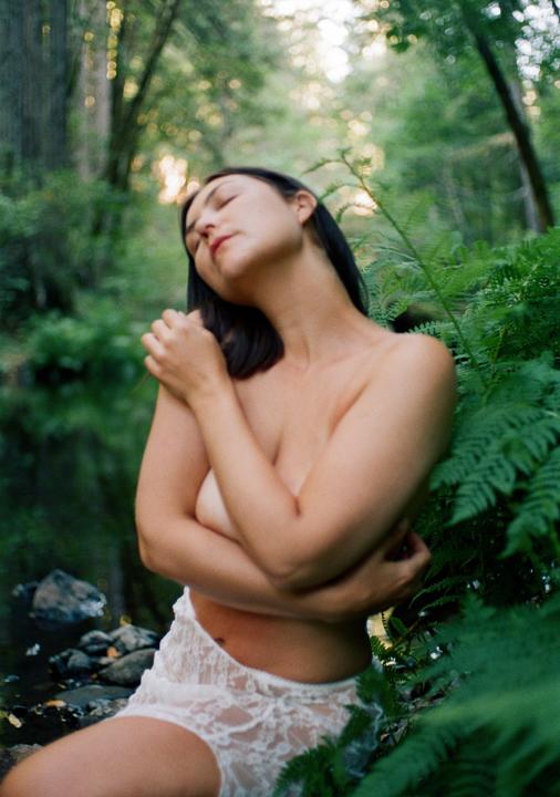 A woman with her eyes closed and arms crossed over her chest is surrounded by lush green foliage and a creek.