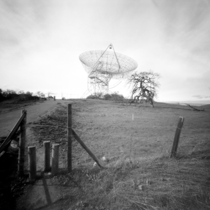 A large satellite dish stands on a hill next to a gnarled tree, with a dirt road and fence in the foreground.