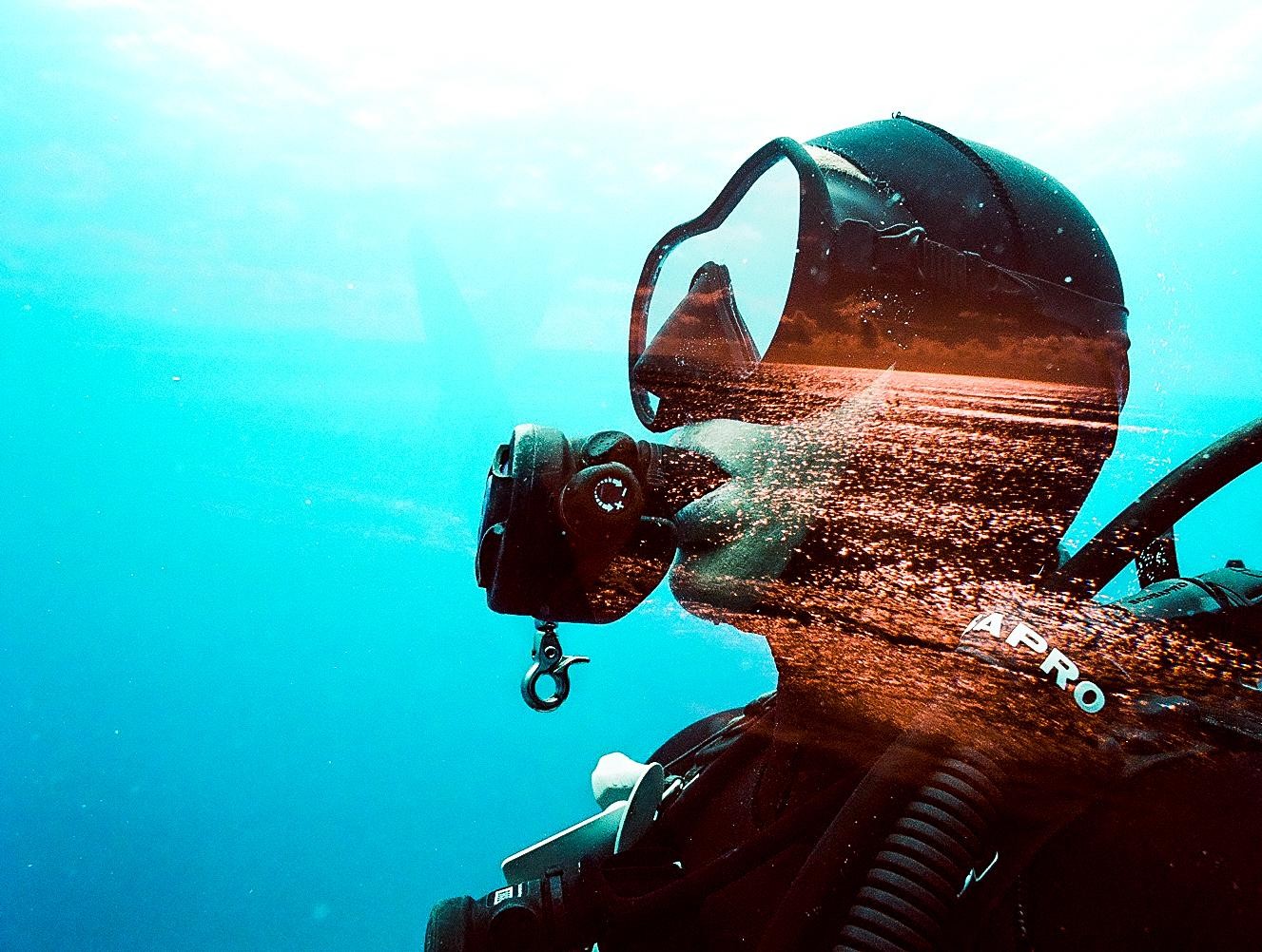 A double exposure shows a diver underwater and a body of water reflecting an orange sky.
