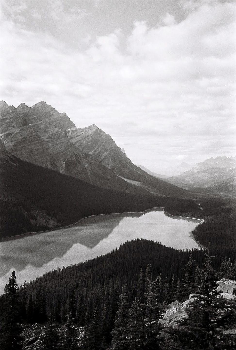 A black and white landscape shows a lake surrounded by mountains and forest under a cloudy sky.