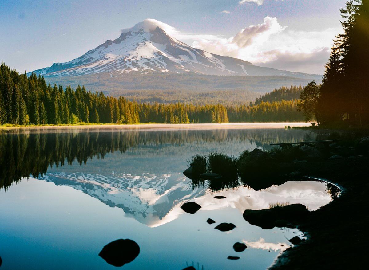 A snow-capped mountain with a cloud on its peak is reflected in a still lake surrounded by a forest.