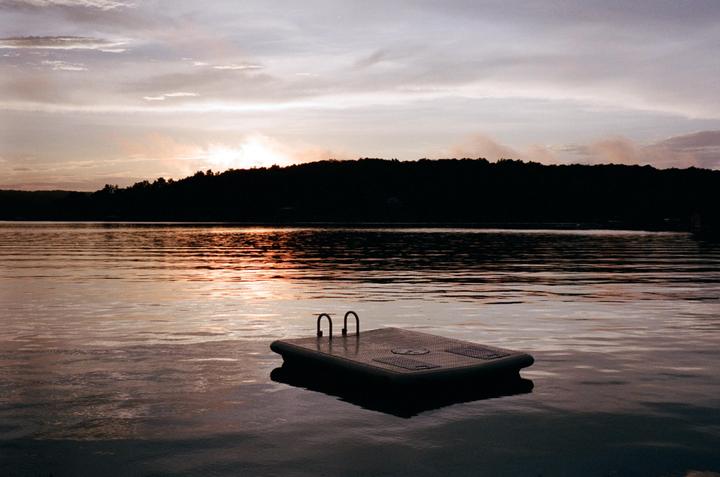 A floating dock sits on a lake with a silhouetted tree line and a colorful sky at dusk.