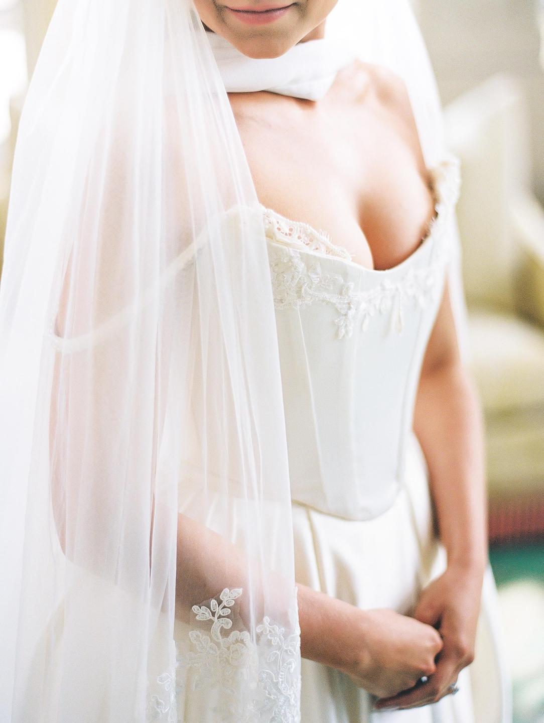 A woman in a white wedding dress and veil stands with her hands clasped.